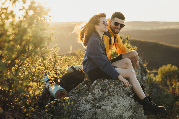 Happy Traveler Couple Man and Woman Sitting on Edge of Rock During Sunset and Smiling, Wind Blowing Womans Hair - Foto, imagen