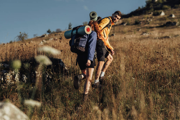 Két utazó hátizsákkal és Camping Mat egy útvonalon, Férfi segít a nőnek előre haladni a hegy tetején naplemente alatt - Fotó, kép