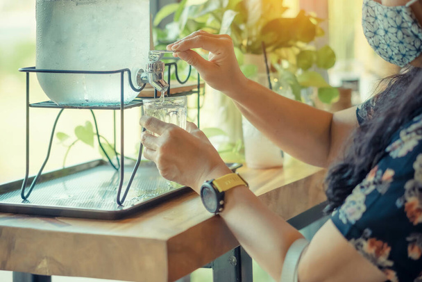Hand der durstigen Frau mit Gesichtsmaske drückt das kühle Trinkwasser in ein klares Glas. Der weibliche Finger drückt einen Knopf kaltes Trinkwasser mit Kühlspender. Gesundheitskonzept. Selektiver Fokus. - Foto, Bild