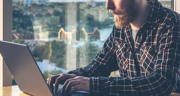 Un hombre con barba trabaja en una computadora, sentado sobre una ventana con vistas a la ciudad, el concepto de TI, freelancing y trabajo remoto. - Foto, Imagen