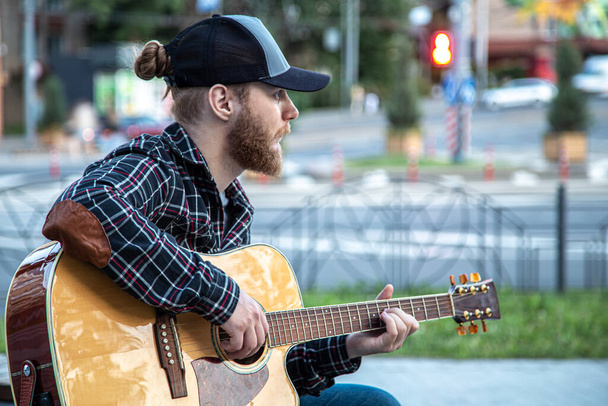 A man street musician with a beard and a cap plays the acoustic guitar. - Foto, Imagen