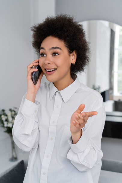 vreugdevolle Afrikaanse Amerikaanse vrouw praten op mobiele telefoon en gebaren in de moderne woonkamer - Foto, afbeelding