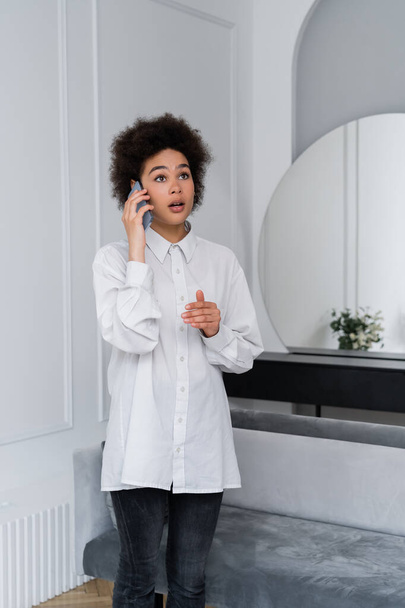 femme afro-américaine choquée parlant sur téléphone portable dans le salon moderne - Photo, image