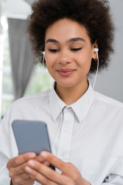 mujer afroamericana sonriente escuchando música en auriculares con cable y usando un teléfono inteligente  - Foto, imagen