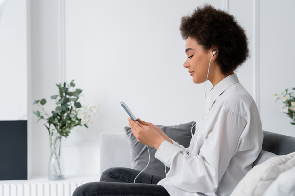 vue latérale de femme afro-américaine heureuse écoutant de la musique et utilisant un smartphone dans le salon - Photo, image