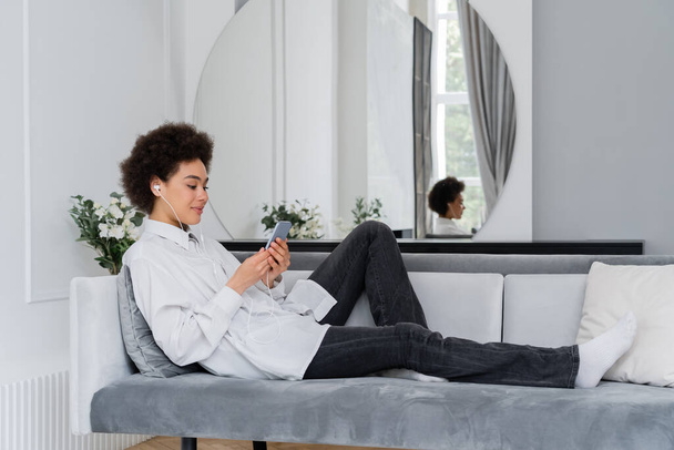 joyful african american woman listening music in wired earphones and using smartphone in modern living room - Photo, image