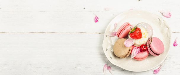 Makarónit eperrel, cseresznyével és bazsarózsával, virágszirmokkal, fehér fa alapon. Édes desszert, romantikus stílus, lapos fektetés, kemény fény, sötét árnyék, banner formátum - Fotó, kép