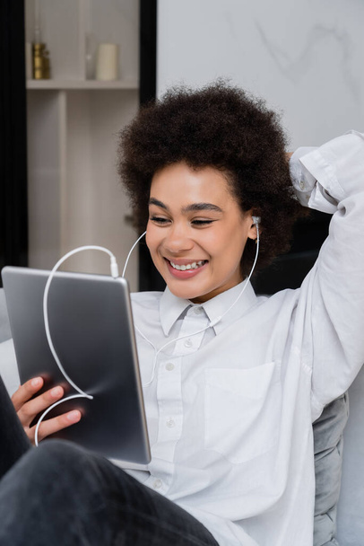 χαρούμενη Αφροαμερικάνα γυναίκα σε ενσύρματα ακουστικά κρατώντας ψηφιακό tablet, ενώ βλέποντας κωμωδία ταινία - Φωτογραφία, εικόνα