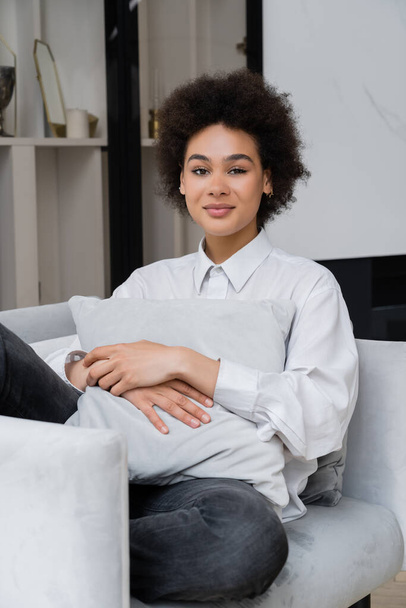 smiling and curly african american woman in white shirt with collar sitting with pillow on grey armchair   - Fotografie, Obrázek
