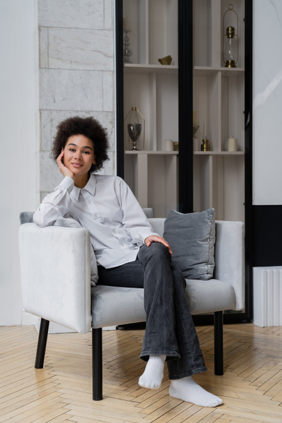 smiling and curly african american woman in white shirt with collar sitting on grey modern armchair   - Fotografie, Obrázek