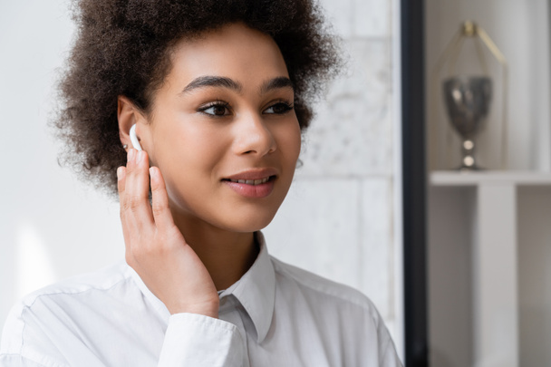 portrait of happy african american woman listening music and adjusting wireless earphone  - Фото, изображение