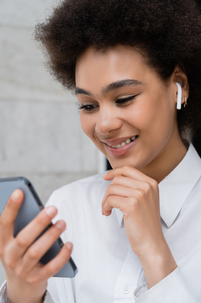 primer plano de la mujer afroamericana feliz escuchando música en auriculares inalámbricos y utilizando el teléfono inteligente - Foto, Imagen