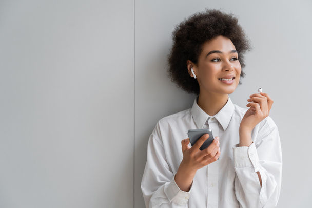 fröhliche afrikanisch-amerikanische Frau hört Musik in drahtlosem Kopfhörer und hält Smartphone in der Nähe der grauen Wand - Foto, Bild