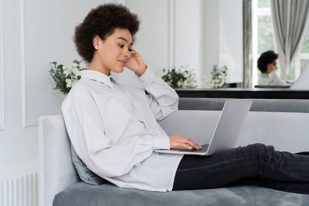 african american woman using laptop while sitting on grey velvet sofa and working from home - Photo, Image