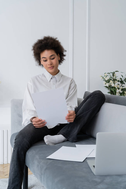 curly african american woman looking at blank document near laptop on velvet grey sofa - Fotografie, Obrázek