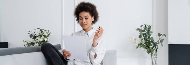curly african american woman holding pen and blank document while working from home, banner - Foto, afbeelding
