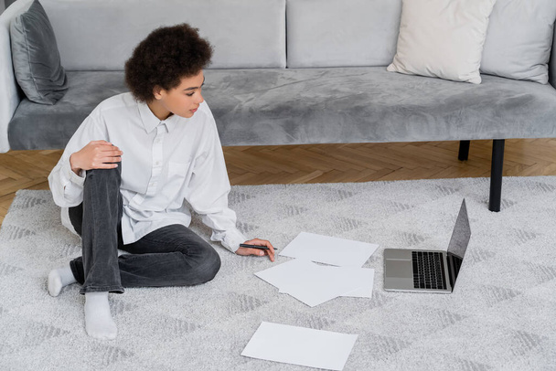 zijaanzicht van Afrikaanse Amerikaanse vrouw op zoek naar laptop terwijl zitten op tapijt in de buurt van documenten - Foto, afbeelding