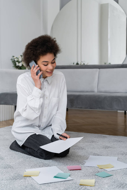 mujer afroamericana hablando en teléfono inteligente mientras mira el documento cerca de papeles en blanco en la alfombra  - Foto, Imagen