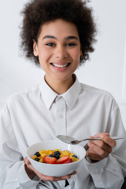 cheerful african american woman holding bowl with corn flakes and berries - Фото, изображение