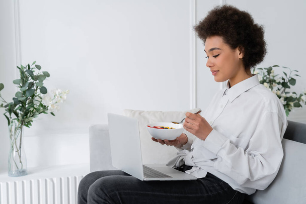 Αφροαμερικάνα γυναίκα κρατώντας μπολ με μούρα, ενώ βλέποντας ταινία στο laptop και κάθεται βελούδινο καναπέ - Φωτογραφία, εικόνα