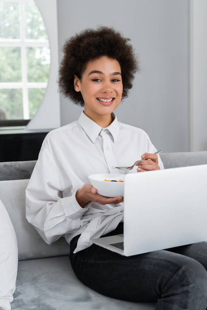 mujer afroamericana feliz sosteniendo tazón con el desayuno mientras mira a la cámara y sentado con el ordenador portátil en el sofá de terciopelo - Foto, Imagen
