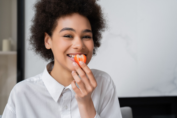 feliz afroamericana mujer comiendo fresa fresca y mirando a la cámara - Foto, Imagen