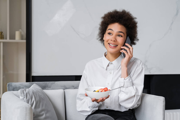 счастливая африканская американка держит миску с завтраком и разговаривает по смартфону - Фото, изображение