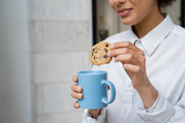 cropped view of happy african american woman holding cup of milk and cookie with chocolate chips  - Fotoğraf, Görsel