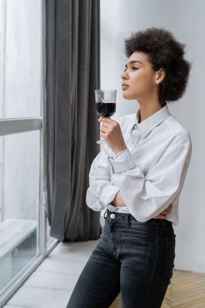 θλιμμένη Αφροαμερικανή που κρατάει ένα ποτήρι κόκκινο κρασί και κοιτάει το παράθυρο. - Φωτογραφία, εικόνα