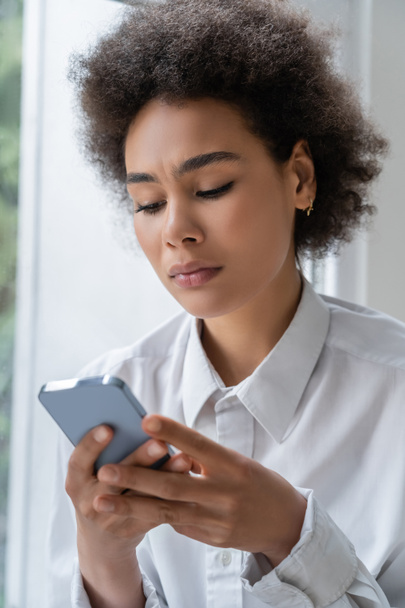 Beyaz tişörtlü üzgün Afrikalı Amerikalı kadın akıllı telefondan mesaj okuyor.  - Fotoğraf, Görsel