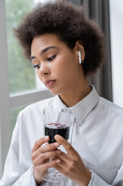καταθλιπτική Αφροαμερικανή γυναίκα με λευκό πουκάμισο και ασύρματο ακουστικό κρατώντας ένα ποτήρι κόκκινο κρασί  - Φωτογραφία, εικόνα