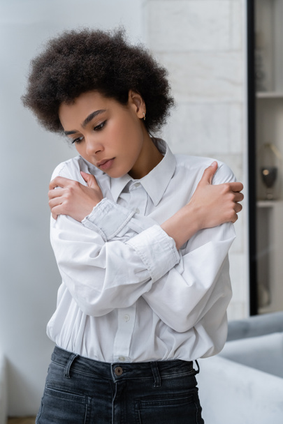 depressed african american woman in white shirt hugging shoulders at home - Фото, изображение