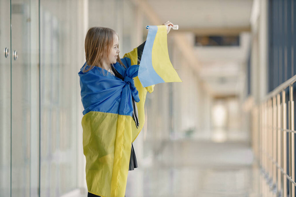 Little girl with ukrainian flag posing for a photo. Young pretty girl standing in front of glass wall. Girl supporting Ukraine. - Foto, Bild