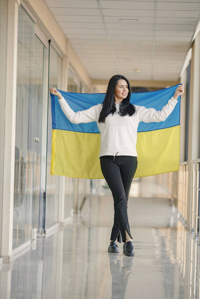 Woman with ukrainian flag posing for a photo. Young pretty girl standing in front of glass wall. Brunette woman wearing black clothes and support Ukraine. - Фото, изображение