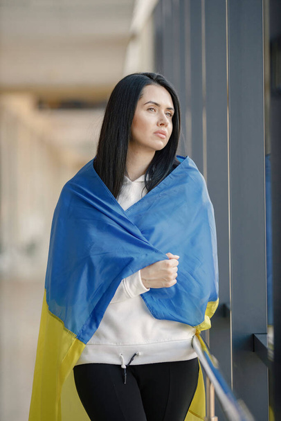 Ukrayna bayrağı taşıyan bir kadın fotoğraf için poz veriyor. Cam duvarın önünde duran genç ve güzel bir kız. Siyah elbiseli esmer kadın Ukrayna 'yı destekliyor.. - Fotoğraf, Görsel