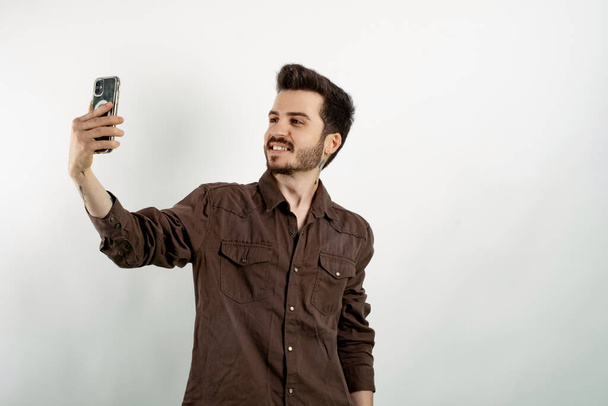 Cheerful caucasian man wearing brown shirt posing isolated over white background making a selfie looking at the phone screen with big smiles. - Zdjęcie, obraz