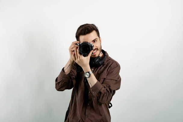 Homem casual feliz vestindo roupas casuais posando isolado sobre fundo branco tirando imagens com câmera dslr. Fotógrafo cobrindo seu rosto com a câmera. - Foto, Imagem