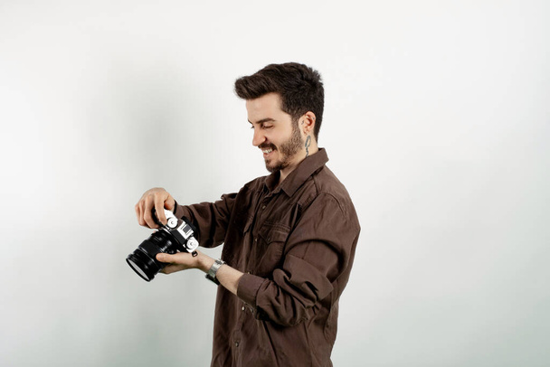 Junger Mann lächelt selbstbewusst in lässiger Kleidung, posiert isoliert vor weißem Hintergrund, hält eine Fotokamera in der Hand und blickt auf den Kamerabildschirm. - Foto, Bild