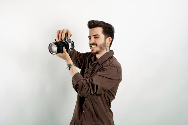 Счастливый случайный человек в повседневной одежде позирует изолированно на белом фоне держа фотокамеру и глядя на экран камеры. - Фото, изображение