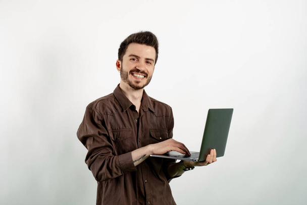 Cheerful young man wearing brown shirt posing isolated over white background looking at the camera and using computer laptop smiling with a happy and cool smile on face. Showing teeth. - Фото, изображение