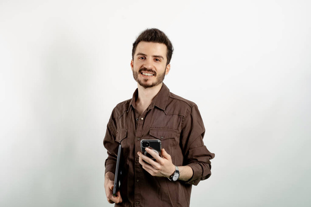 Portrait of young handsome man wearing brown shirt posing isolated over white background holding in hands carrying laptop and phone posing. Looking at the camera with big smiles. - Photo, image