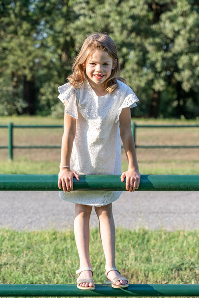 cute little blonde girl with white dress above an iron fence outdoors - grass and plants in a park in the background - Photo, image