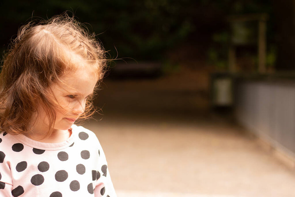 Μικρό λυπημένο κοκκινομάλλικο κοριτσάκι με φόρεμα κοιτάζει κάτω. παιδί στο πάρκο - Φωτογραφία, εικόνα