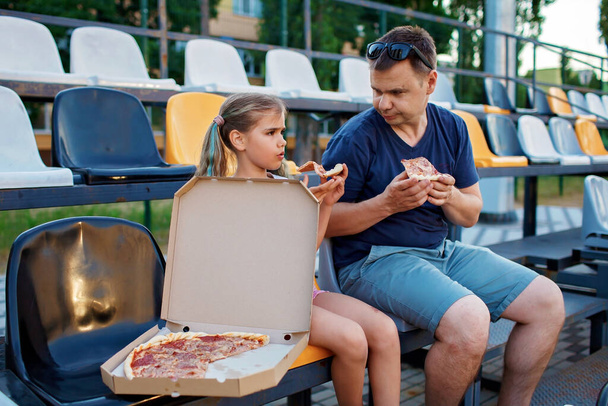 Père avec sa fille, fans de football, regarder le match de football, manger de la pizza et encourager l'équipe locale au stade, émotions réelles, événement sportif et soutien des fans, style de vie en plein air, nourriture de rue - Photo, image