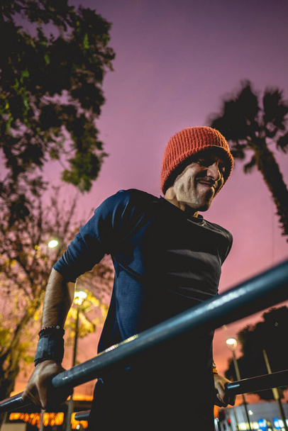 Fit and handsome athletic latin guy with wool cap doing a calisthenic exercise routine (performs dips on the parallel bars) in an street workout park with beautiful and colorful sunset sky - Photo, Image