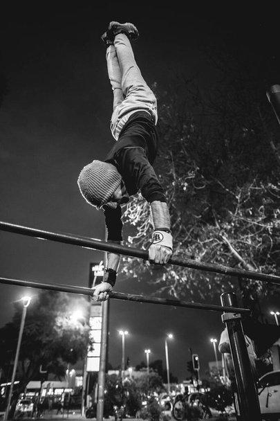 Healthy and fit young latin man with wool cap and wristbands doing calisthenics (inverted pose on high bars) on a street workout park with beautiful and colorful sunset sky (in black and white) - Photo, Image