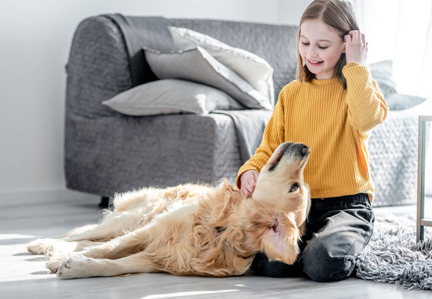 Preteen girl playing with golden retriever dog sitting on floor. Pretty kid child petting doggy pet at home - Valokuva, kuva