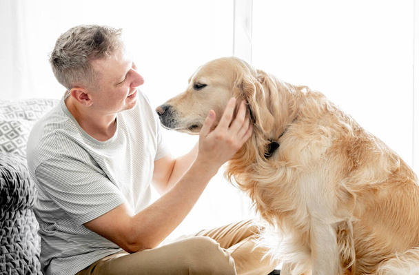 Young handsome man with his golden retriever dog in bright living room. - 写真・画像