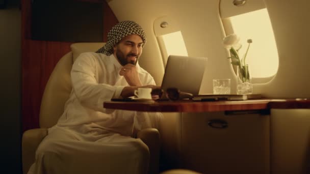 Улыбающийся бизнесмен печатает клавиатуру на самолёте. Веселый мужчина в чате онлайн в деловой поездке. Красивый арабский рабочий компьютер просматривает интернет в мусульманской одежде. Расслабленный менеджер наслаждается полетом - Кадры, видео