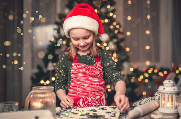 Porträt eines kleinen Mädchens mit Weihnachtsmannhut, das Plätzchen auf dekoriertem Weihnachtsbaum schneidet - Foto, Bild